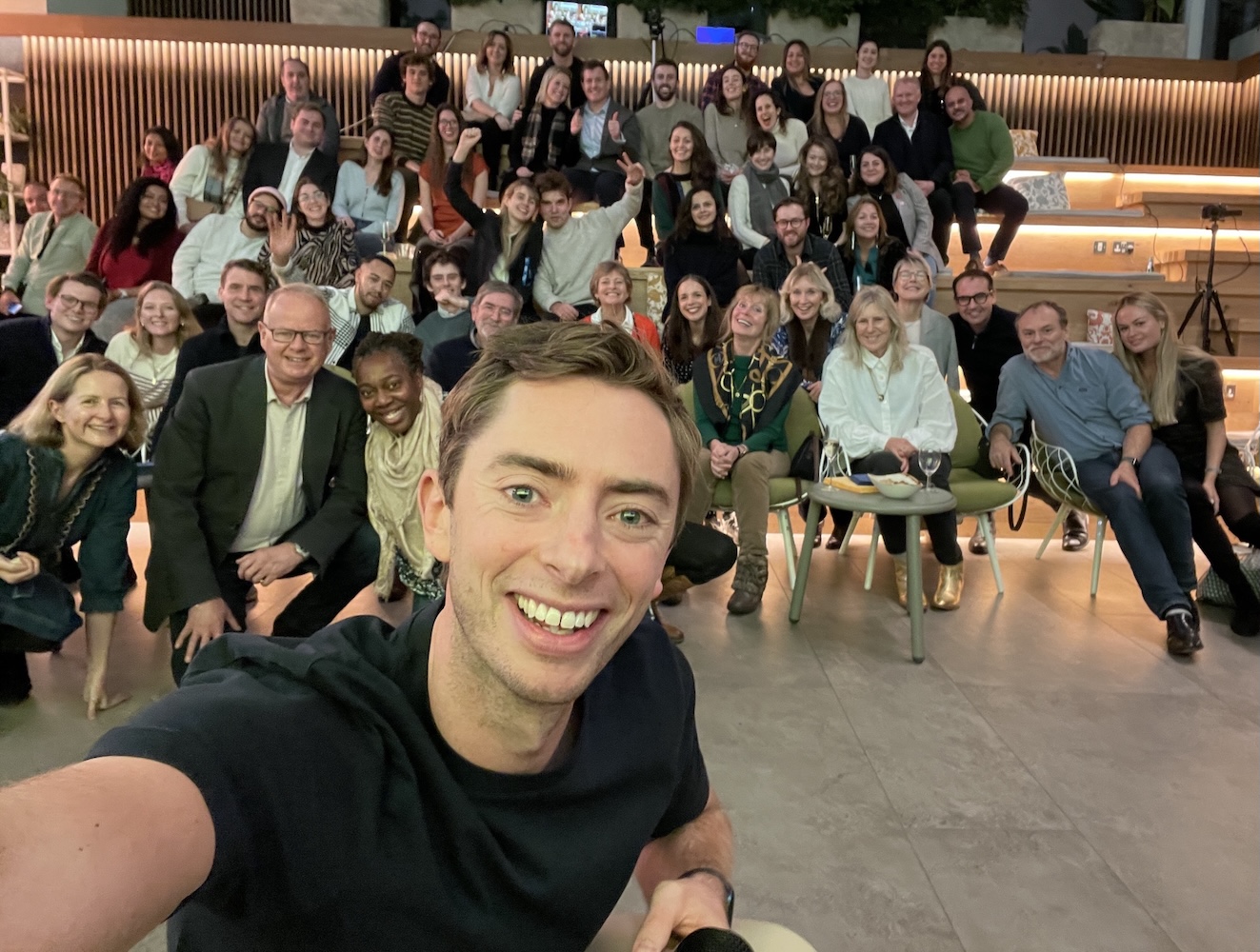 Alex Merry takes a selfie with a large group of attendees at an indoor event